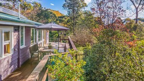 A wooden deck with a table and chairs, a covered seating area, and a view of surrounding trees and hills. - Cooee Cottage @ Blackheath (Blackheath)