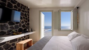 A bedroom with a stone wall, a flat-screen TV, a bed with white linens, and a view of the sea through open doors.