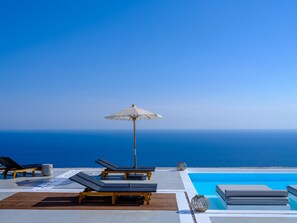 A poolside area with lounge chairs, a pool, and a view of the ocean. - Terra Blanca Suites (Santorini)