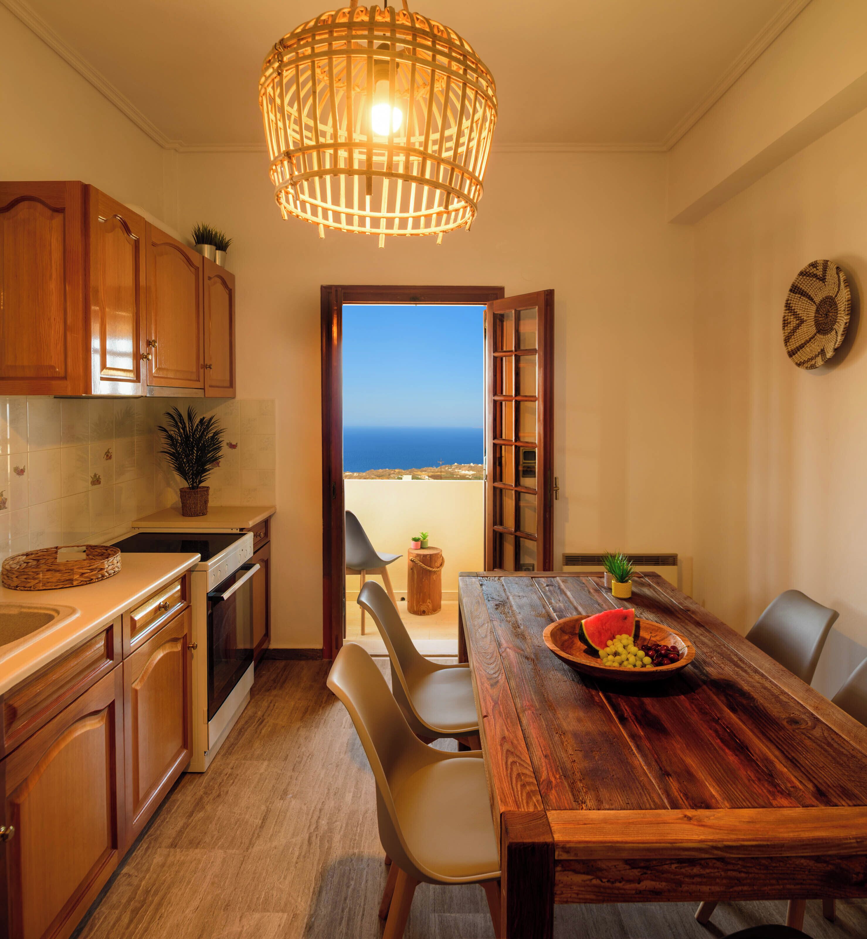 A kitchen with wooden cabinets, a dining table with chairs, a wooden bowl of fruit, and a view of the sea through an open door.