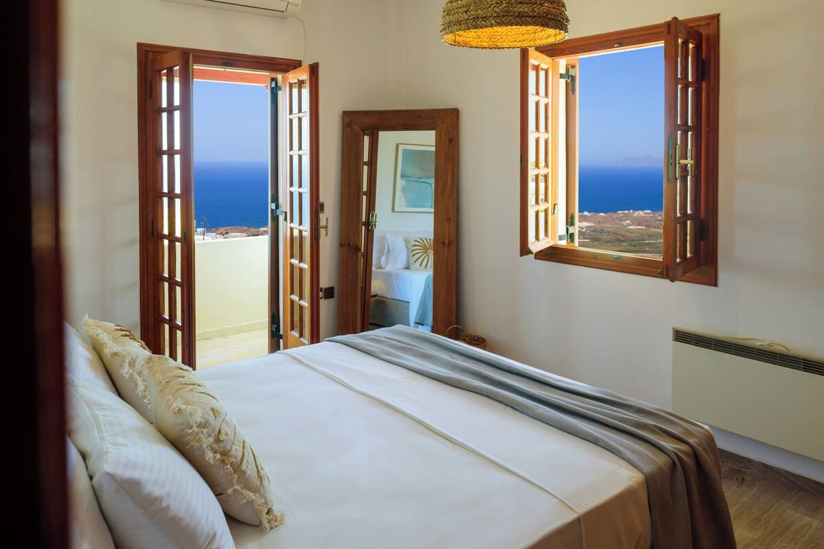 Un dormitorio con cama, vista al mar a través de contraventanas abiertas y una puerta que conduce a otra habitación.