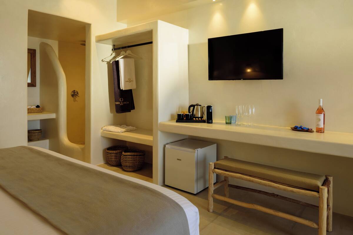 Una habitación de hotel moderna con una cama, un televisor de pantalla plana montado en la pared, una kitchenette pequeña y un banco de madera.