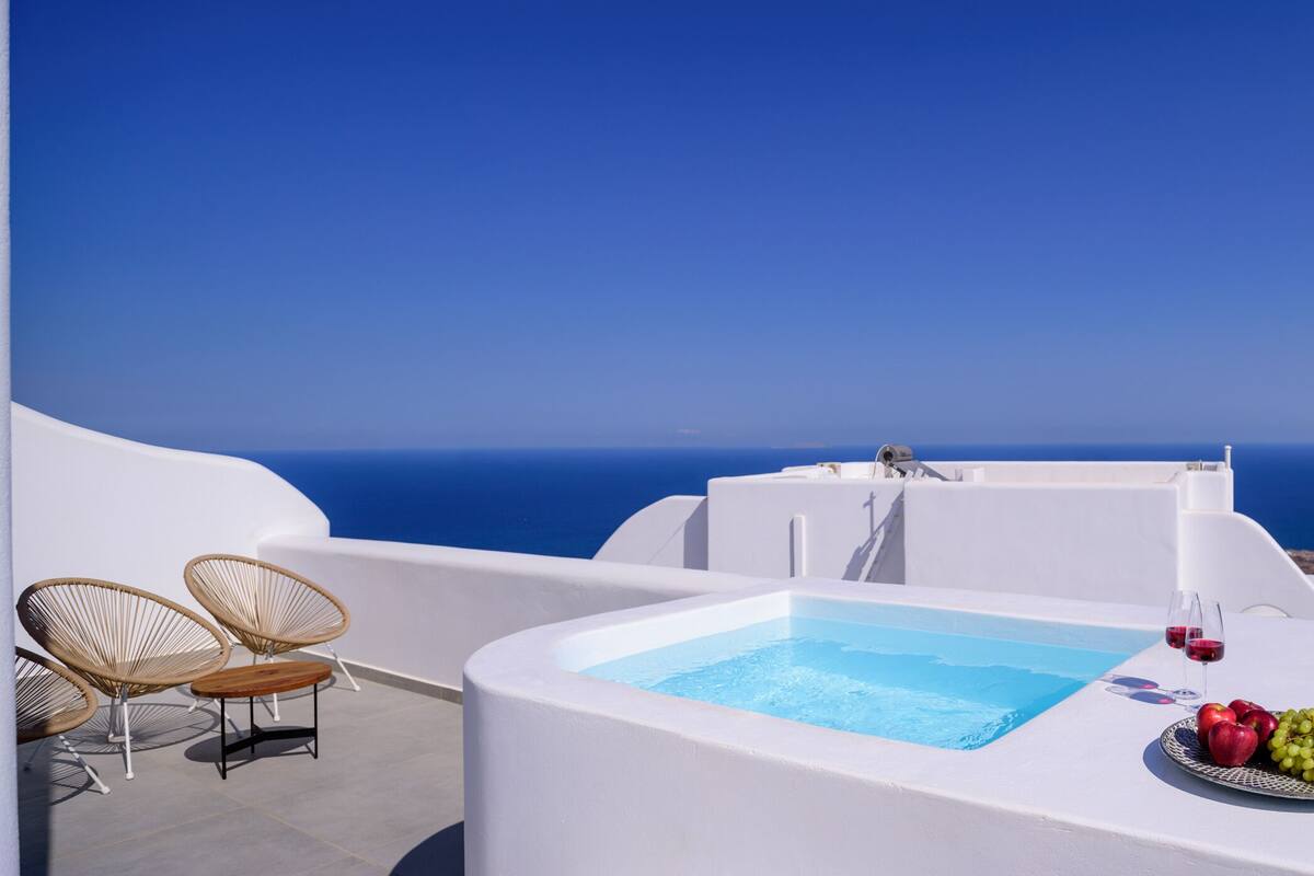 Zona de piscina con vista al mar, mobiliario blanco y una bandeja con fruta y copas de vino.