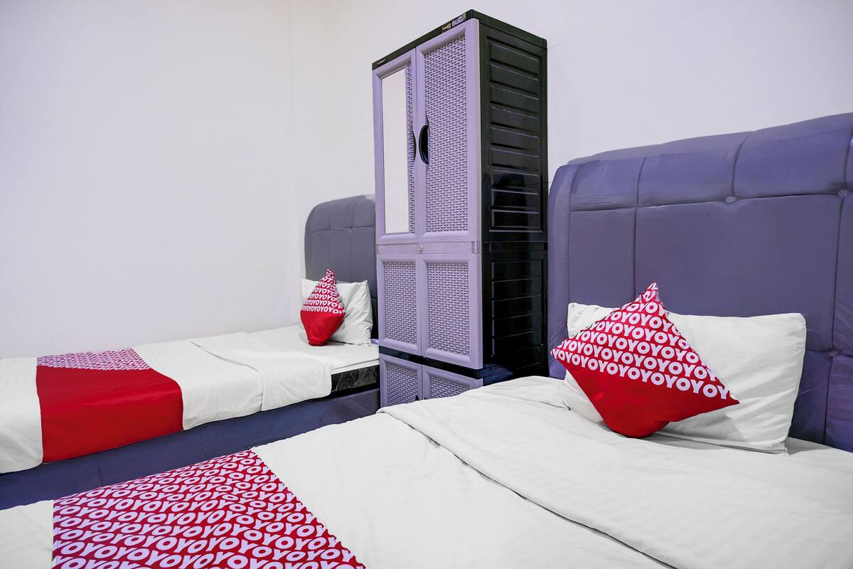 A hotel room with two beds, each with a red and white patterned pillow, and a large wardrobe in the center.