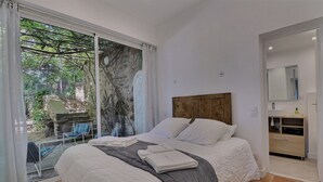 A bedroom with a large bed, a wooden headboard, a bathroom with a mirror and sink, and a view of greenery outside. - Apartment, 1 bedroom, Shared pool, Private beach, Mediterranean garden (La Croix-Valmer)