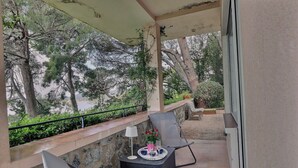 A covered outdoor seating area with a table set for two, a chair, and a view of trees and greenery. - Apartment, 1 bedroom, Shared pool, Private beach, Mediterranean garden (La Croix-Valmer)