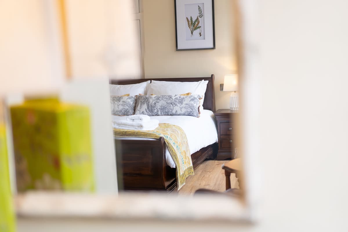 A neatly made bed with a patterned blanket and pillows, a bedside table with a lamp, and a framed botanical print on the wall.