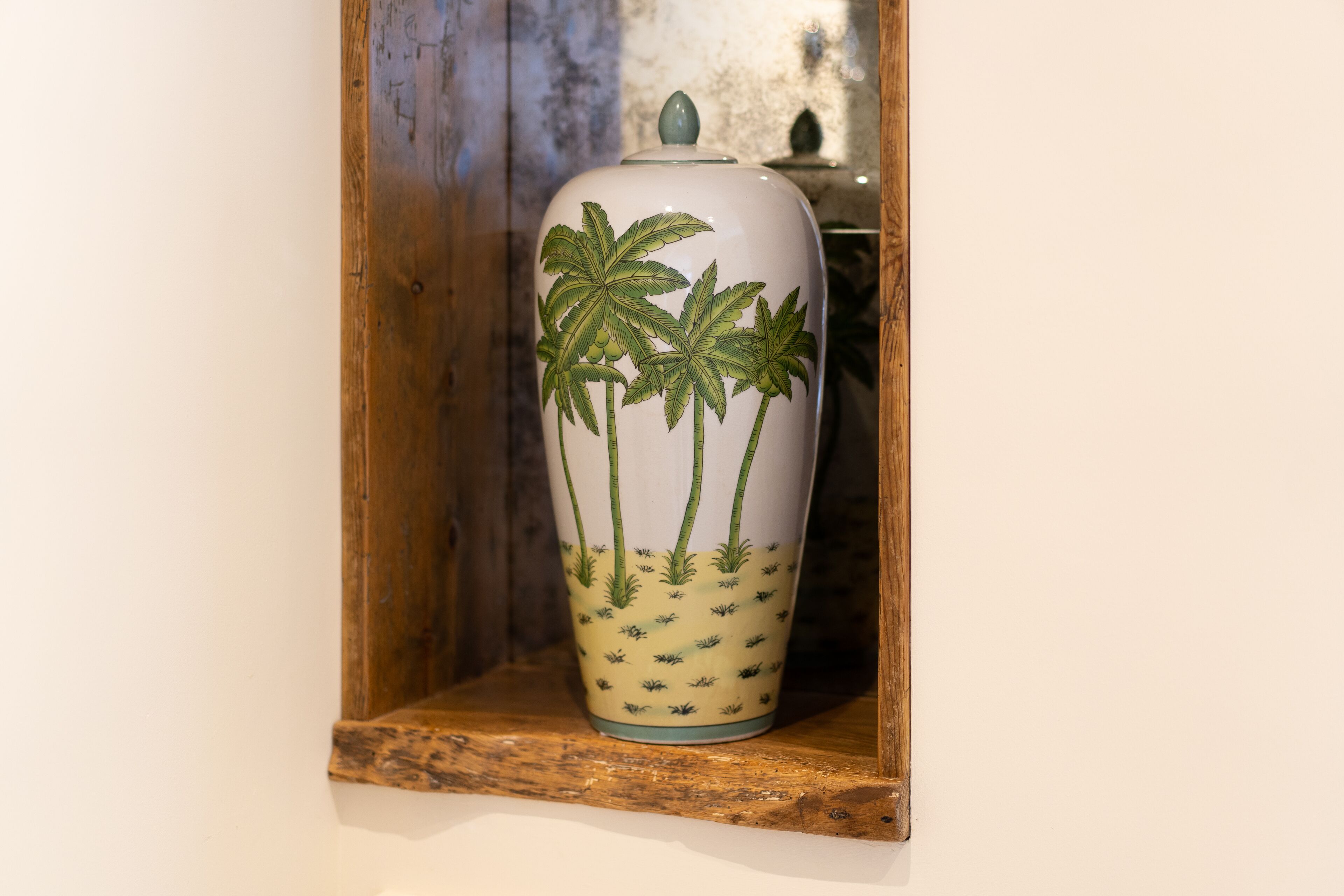 A white ceramic vase with green palm tree designs on a wooden shelf.