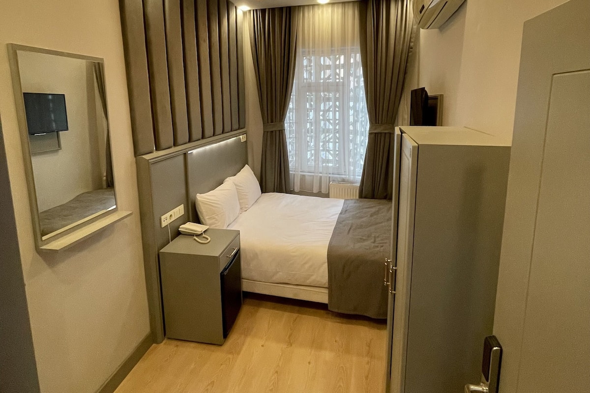 A hotel room with a bed, bedside table, and a mirror. There is a window with curtains, a ceiling light, and an air conditioning unit.