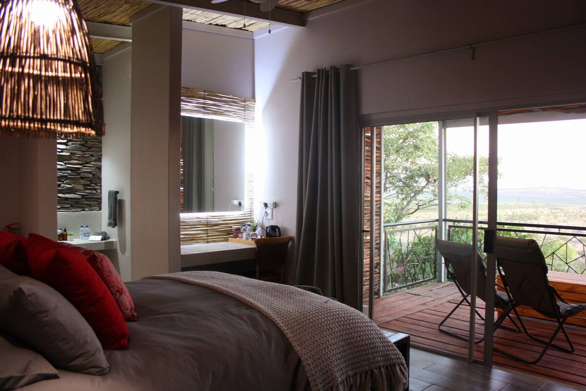 A bedroom with a large bed, a desk, a chair, and a balcony with a view.