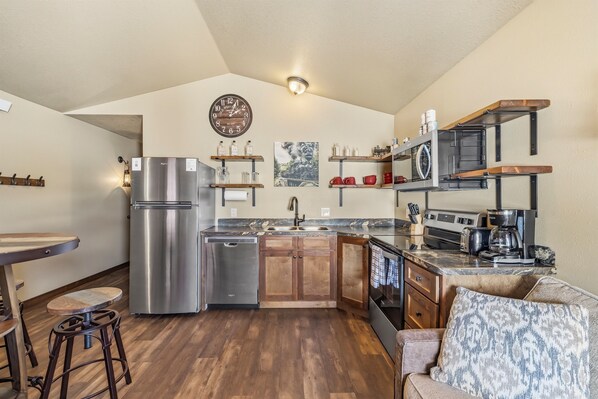 A compact kitchen with a stainless steel refrigerator, wooden cabinets, and a built-in oven. There are bar stools and a small dining table.