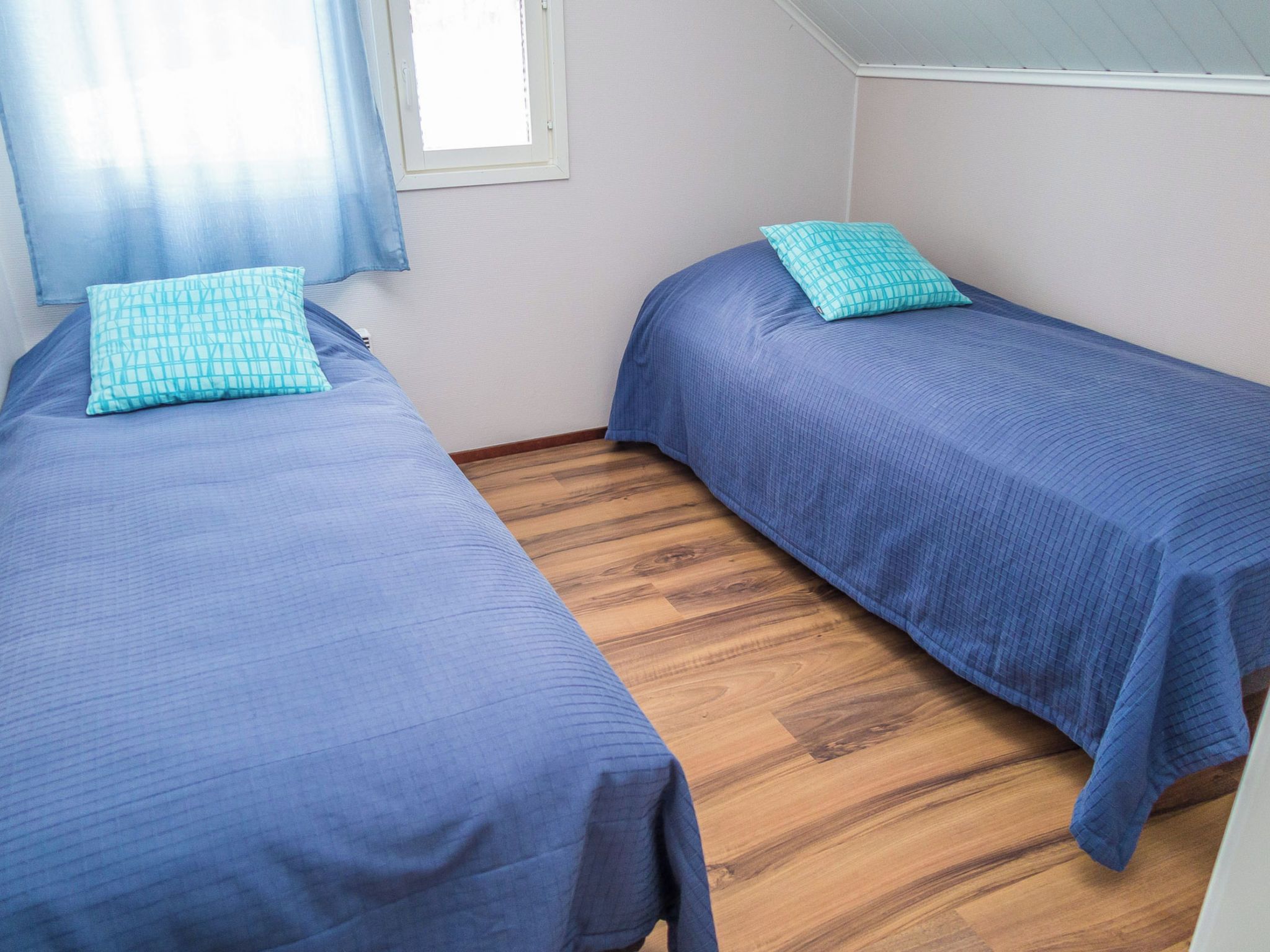 Una habitación con dos camas, cada una con cubrecama azul y almohada a cuadros. El piso es de madera y hay una ventana con marco blanco.