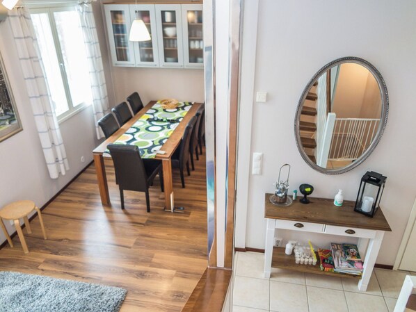 A dining area with a long table, chairs, a small stool, a mirror, a shelf with books, and a window with curtains.