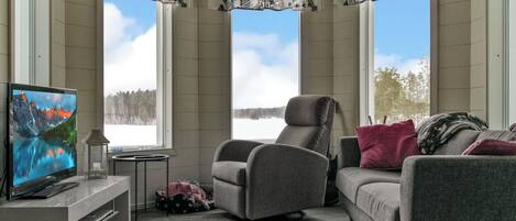 Ein Wohnzimmer mit Fernseher, einem grauen Sessel, einer Couch und Blick auf eine verschneite Landschaft durch große Fenster.