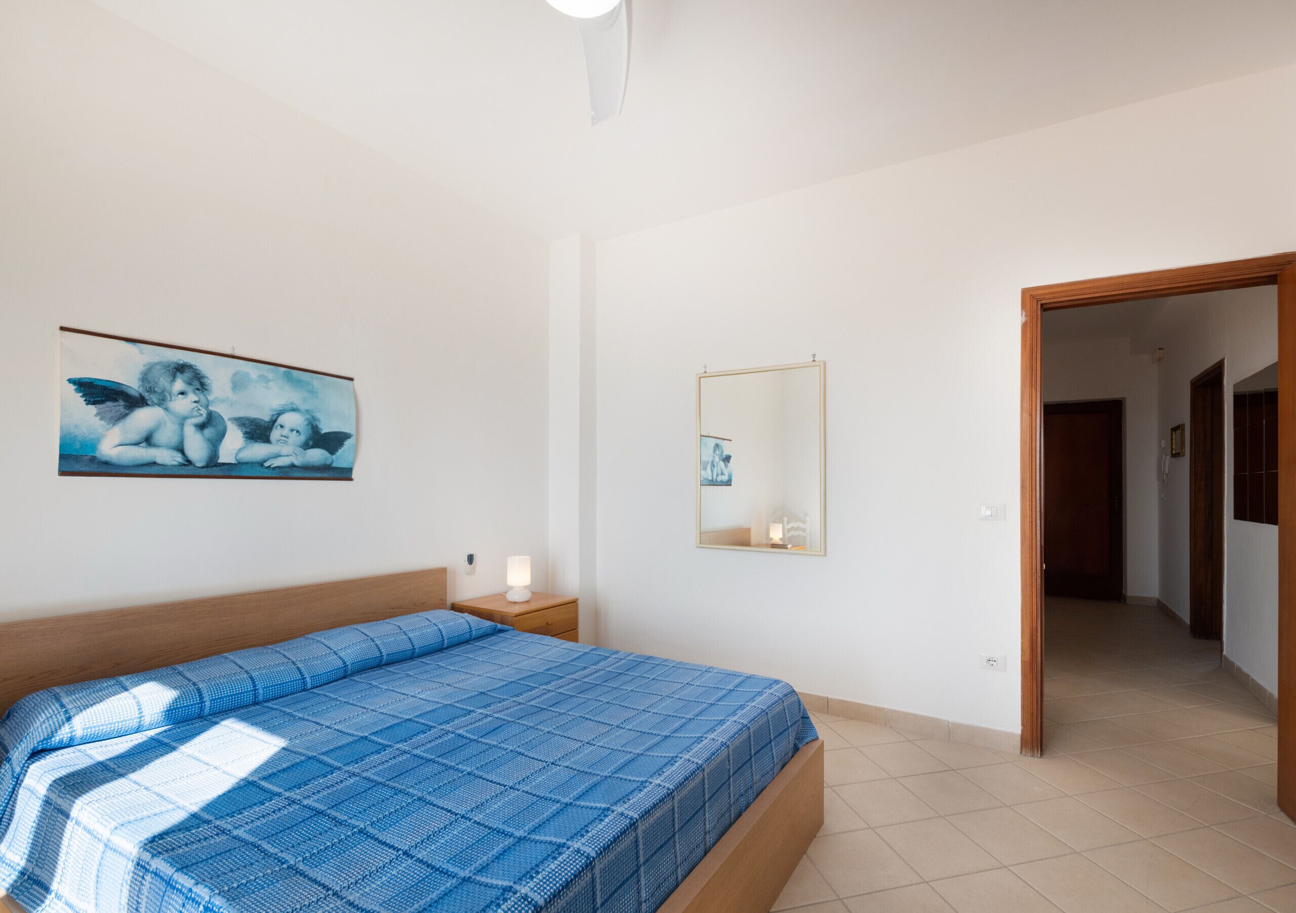 A bedroom with a bed, a nightstand, a wall-mounted mirror, and a door leading to another room.