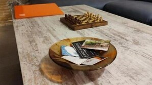 Una stanza con parete in mattoni, un divano, un tavolino da caffè con scacchi e riviste, e una porta con cornice verde.
