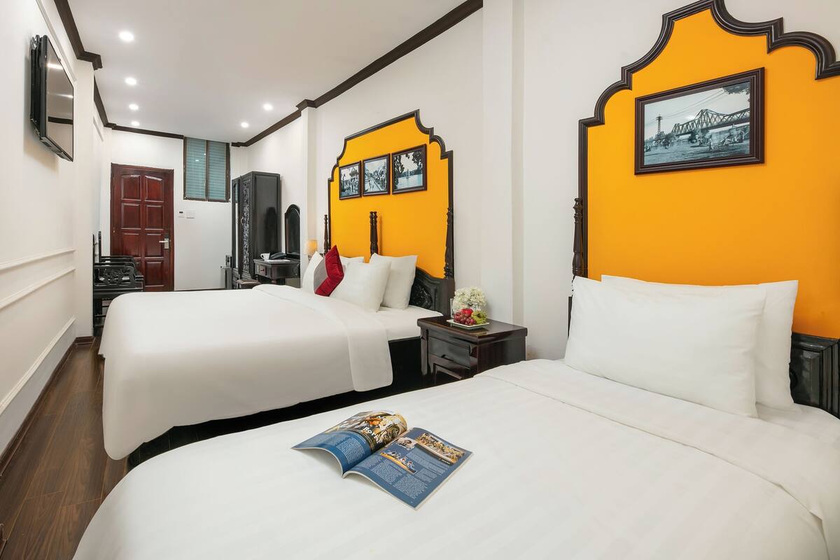 A hotel room with two beds, a wooden wardrobe, a nightstand with a lamp, and a framed picture on the wall.