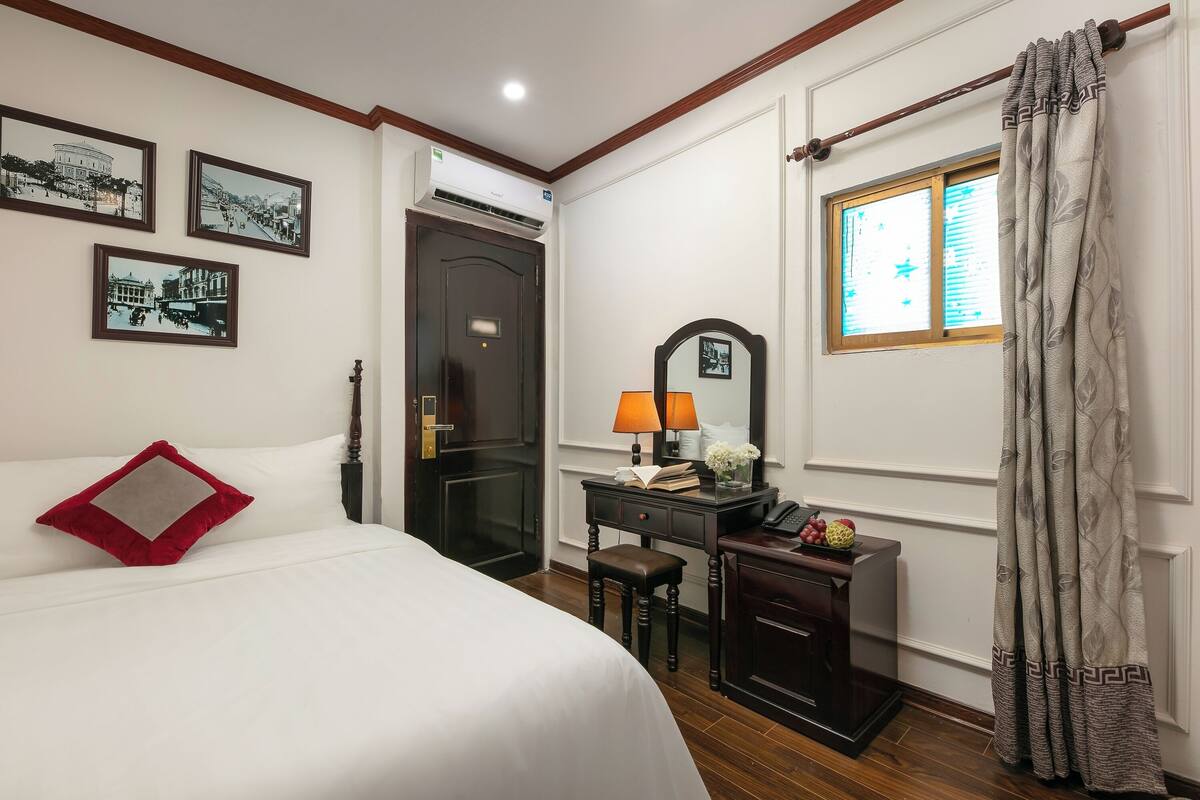 A hotel room with a bed, a desk with a lamp, a mirror, and a window with curtains.