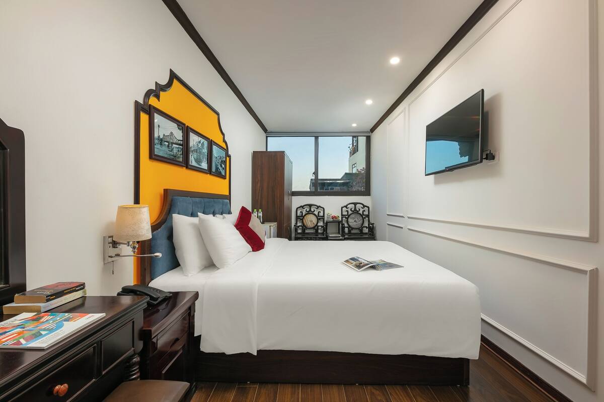A hotel room with a large bed, a bedside table with a lamp, a TV mounted on the wall, and a window with a city view.