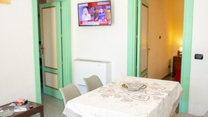 Ein Zimmer mit einem Esstisch, der mit einer Spitzentuch-Tischdecke bedeckt ist, einem an der Wand befestigten Fernseher und einer weißen Klimaanlage.