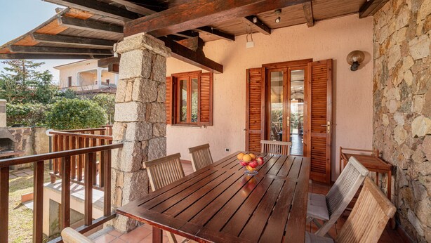 Een houten tafel met stoelen en een fruitschaal, op een balkon met stenen muren en houten luiken.