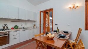 Een kleine keuken met witte kastjes, een houten eettafel en een rieten stoel.