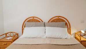 Een tweepersoonsbed met een beige sprei en twee kussens, omringd door rieten nachtkastjes met lampen.
