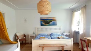 Une chambre à coucher comprenant un lit, des tables de chevet, une chaise, un banc, un luminaire suspendu, un tableau et une fenêtre avec des rideaux.