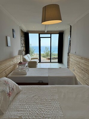 Habitación de hotel con una cama grande, una cama más pequeña, una mesita de noche, una lámpara y vistas al mar a través de una puerta de cristal.