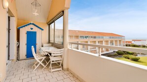 Uma varanda com mesa e cadeiras, uma porta azul e vista para um edifício com telhado vermelho.