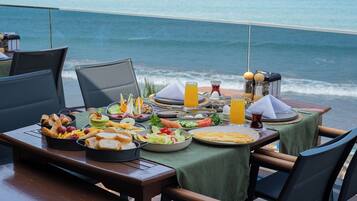 Una mesa con diversos alimentos para el desayuno, como pan, queso y frutas, con vista al mar.