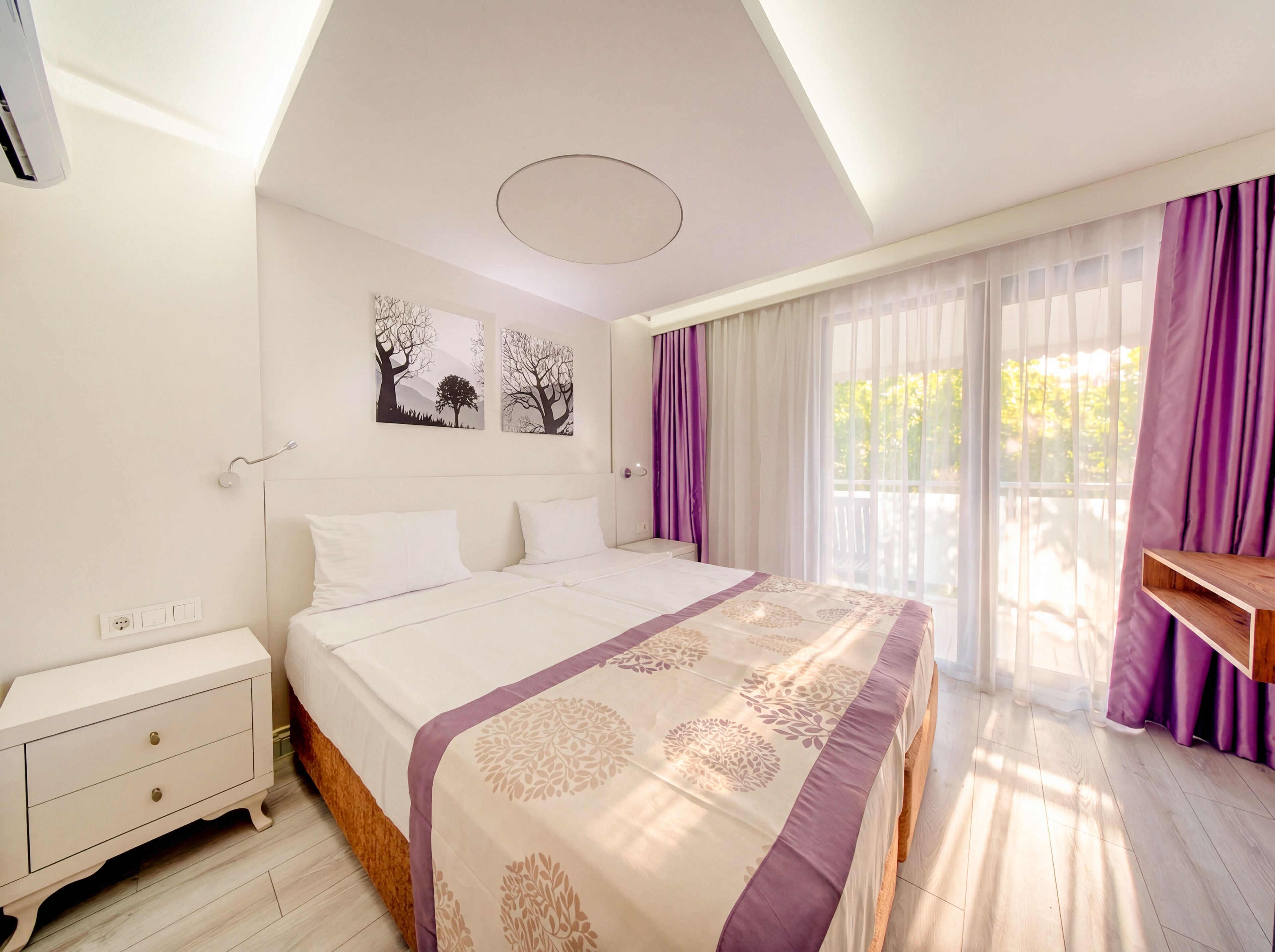 A bedroom with a bed, a nightstand, a wall-mounted light, and a window with curtains.