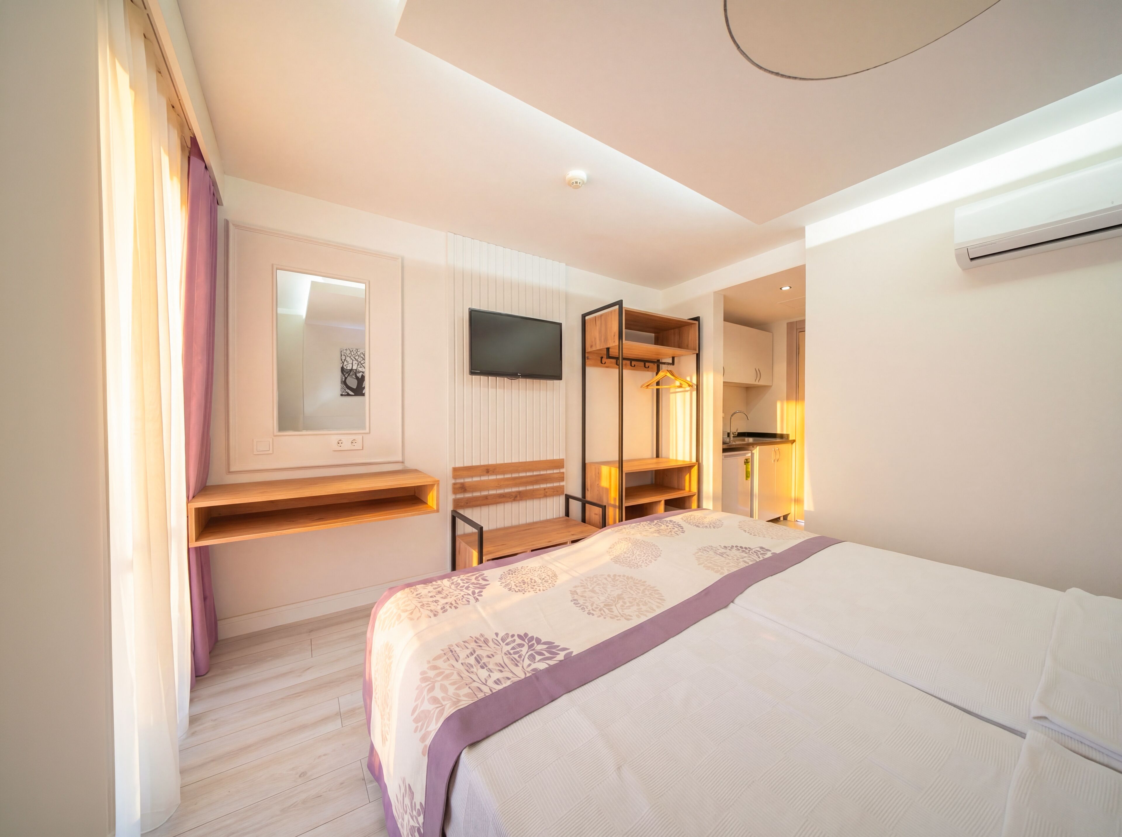 A modern hotel room with a bed, a TV, a mirror, a wooden bench, and a small kitchenette.