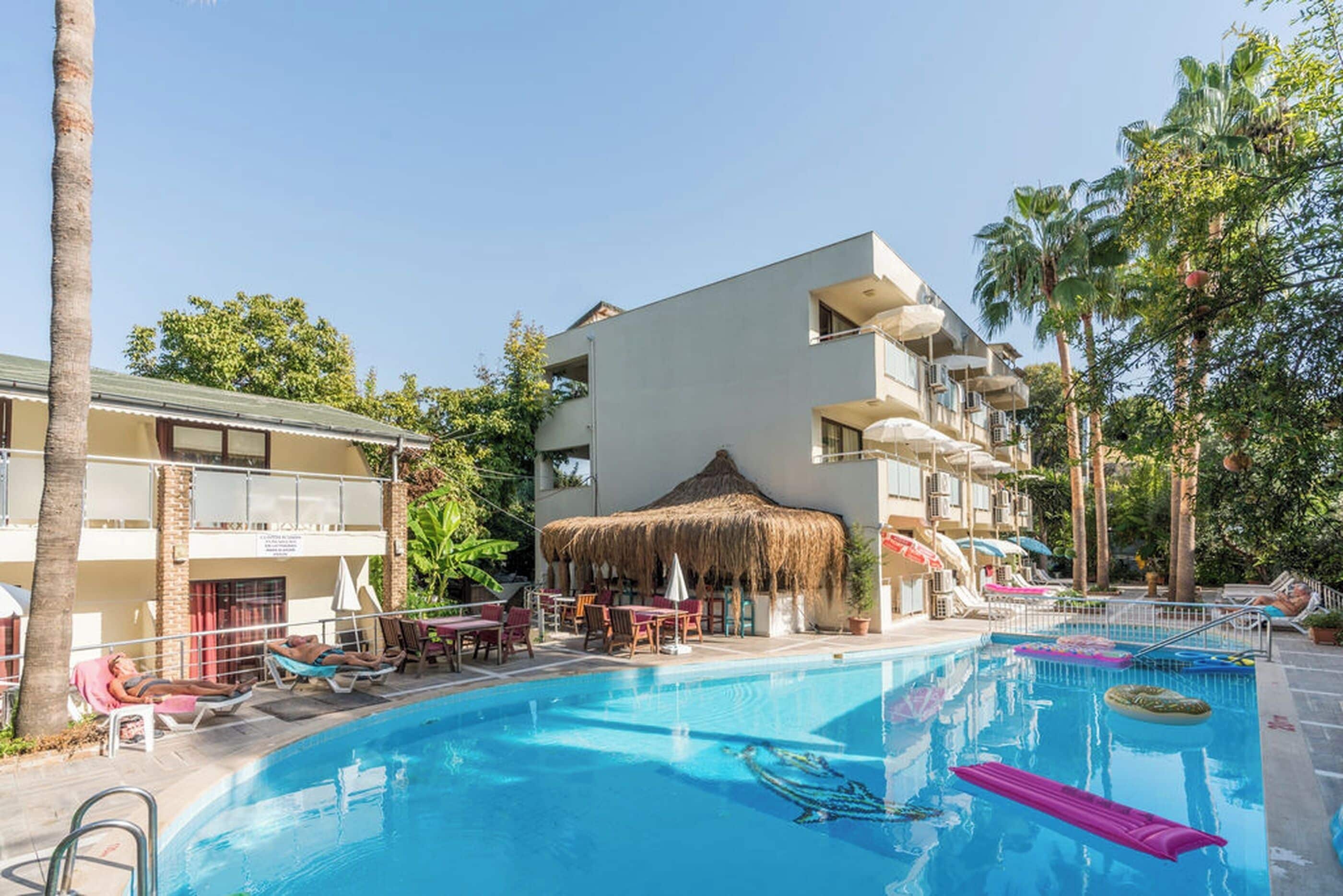 Una piscina con sillas de descanso y sombrillas, rodeada de palmeras y un edificio con balcones.