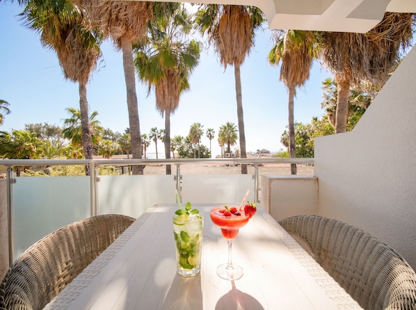 A balcony with wicker furniture, a table with a drink, and a view of palm trees and the beach.