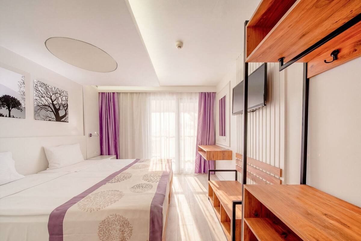 A hotel room with a bed, a wooden bench, a TV mounted on the wall, and a window with curtains.