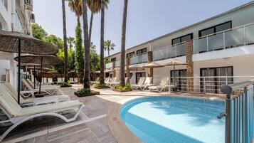 A pool area with lounge chairs and umbrellas, surrounded by palm trees and a building with balconies.