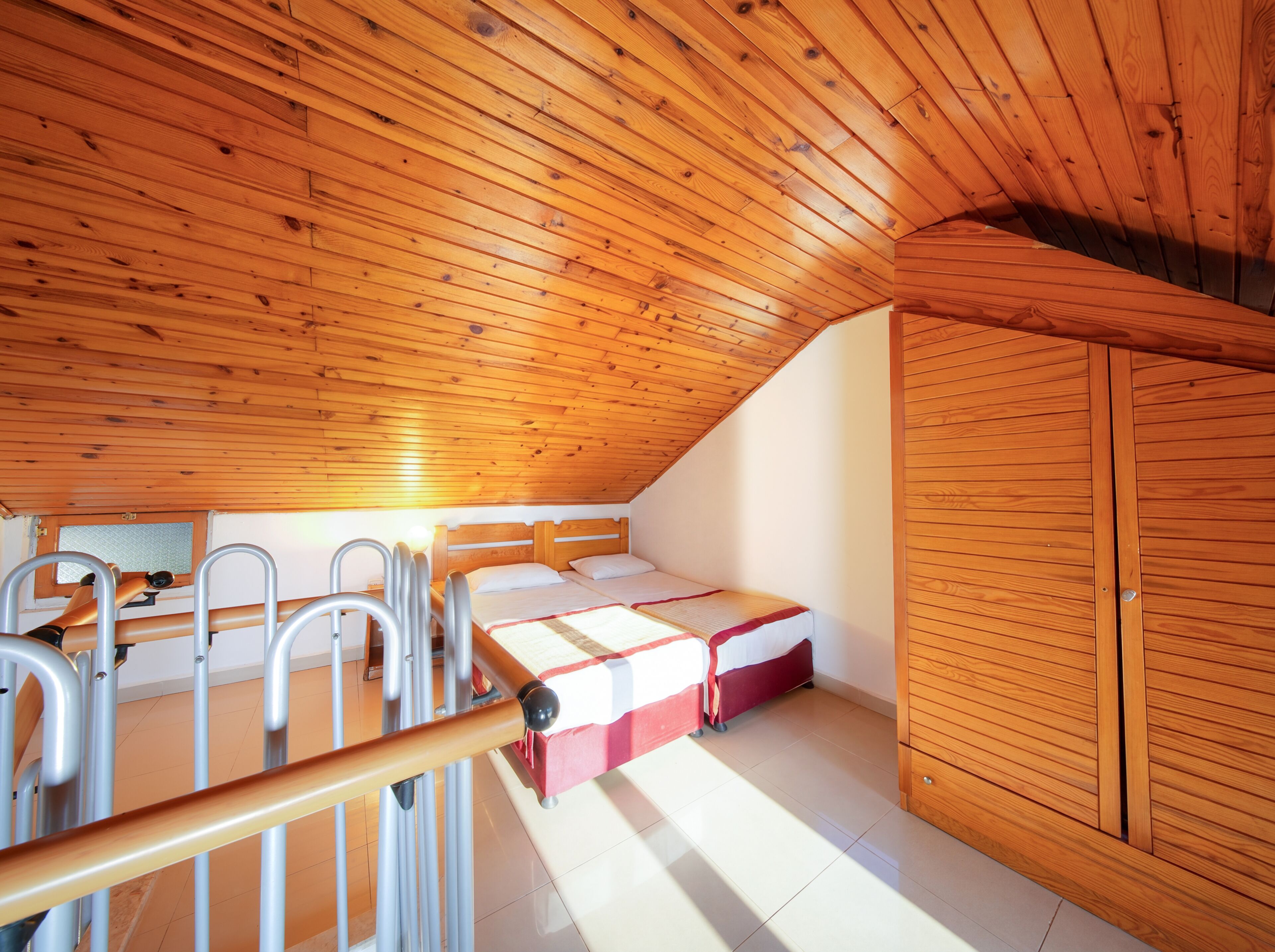 A room with a sloped wooden ceiling, a bed with a red and white bedspread, a wooden wardrobe, and a window with blinds.