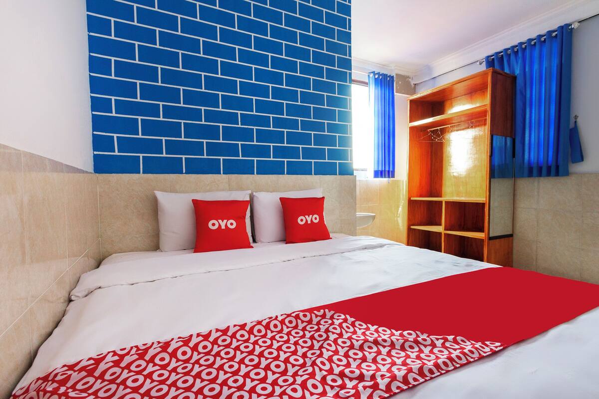 Un lit recouvert d’une couvre-lit à motifs blancs et rouges, deux oreillers ornés du texte « OYO », un mur en briques bleues et une armoire en bois avec des rideaux bleus.