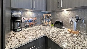 A kitchen with dark wood cabinets, a granite countertop, a coffee maker, jars of condiments, a knife block, a toaster, and a bottle of water. - Apt 335 · Luxury 2Bd Queen Unit | Free Parking |Hwy121| TPC (McKinney)
