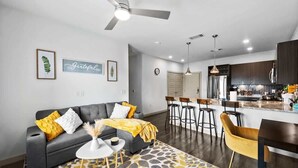 A modern living room with a grey sofa, yellow throw, and a patterned rug. A kitchen with dark cabinets and stainless steel appliances is visible in the background. - Apt 335 · Luxury 2Bd Queen Unit | Free Parking |Hwy121| TPC (McKinney)