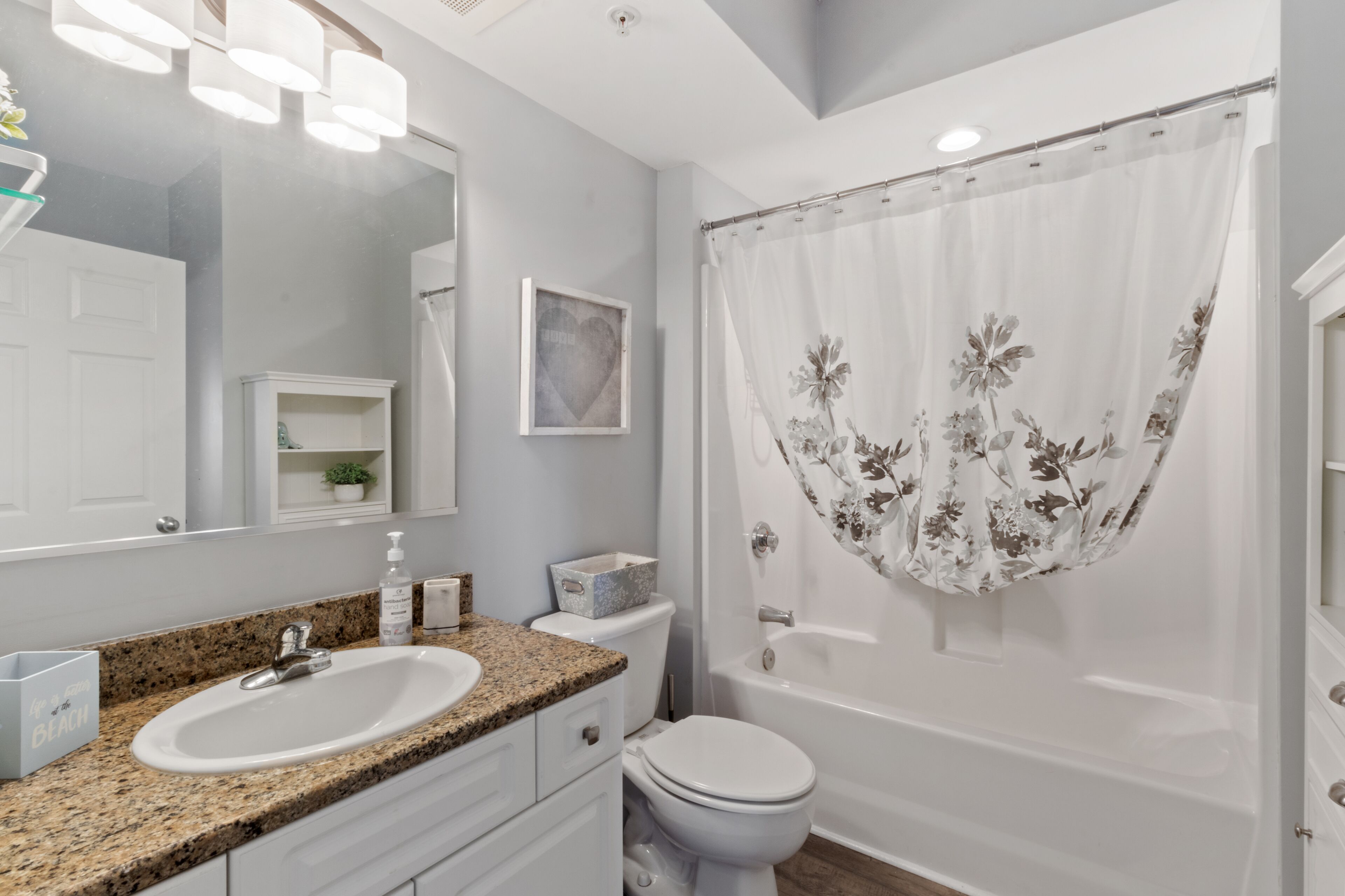 A bathroom with a granite countertop sink, a white toilet, a bathtub with a floral shower curtain, a white cabinet, and a mirror above the sink.