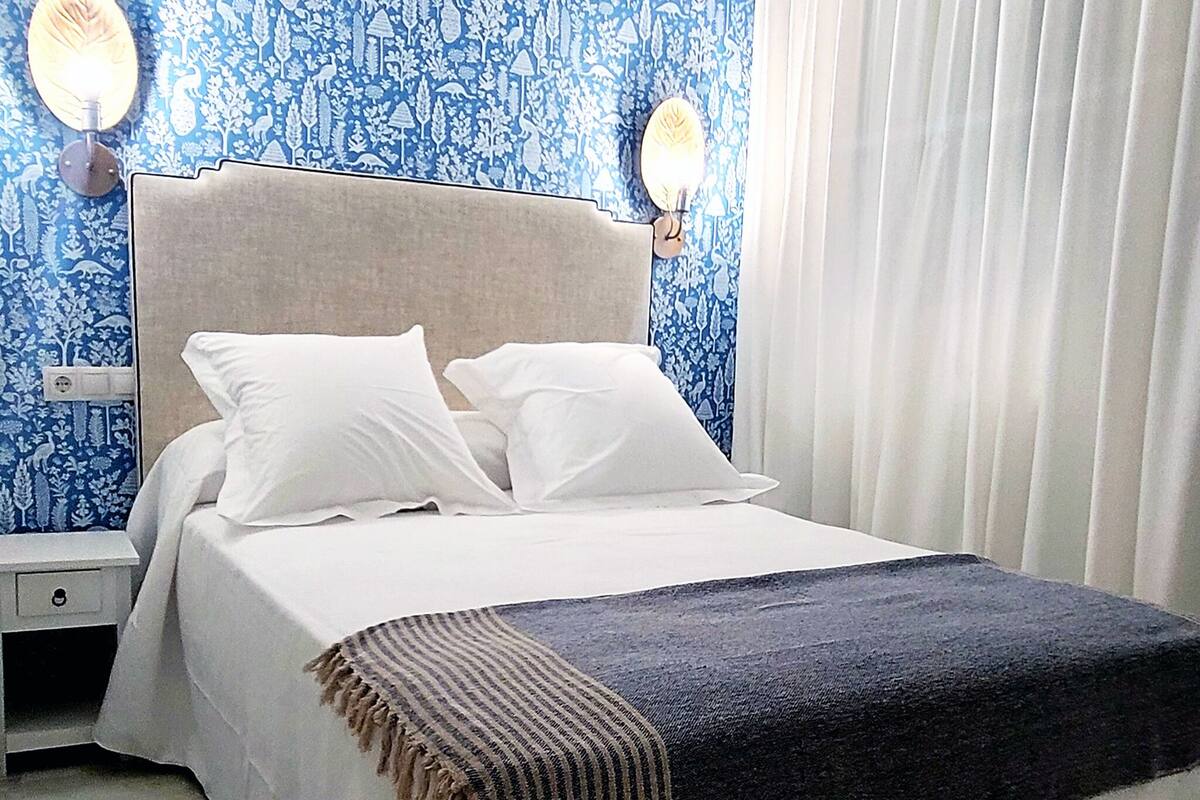 A bedroom with a bed, two bedside lamps, a nightstand, a blue patterned wall, and a white curtain.