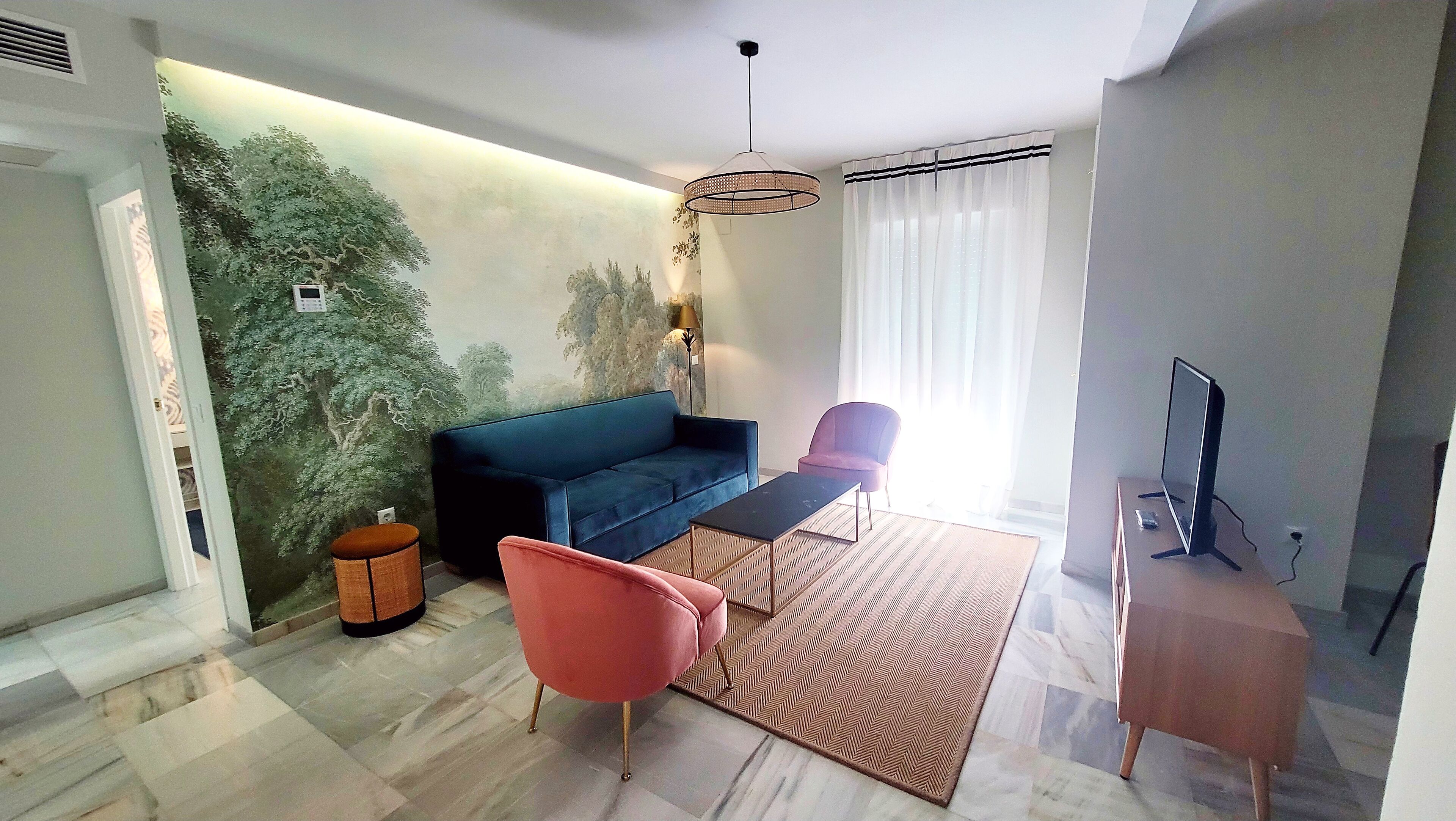 Una sala de estar moderna con un sofá azul, una mesita, un sillón rosa, un televisor sobre un mueble bajo y una pared decorativa con un mural de un árbol.