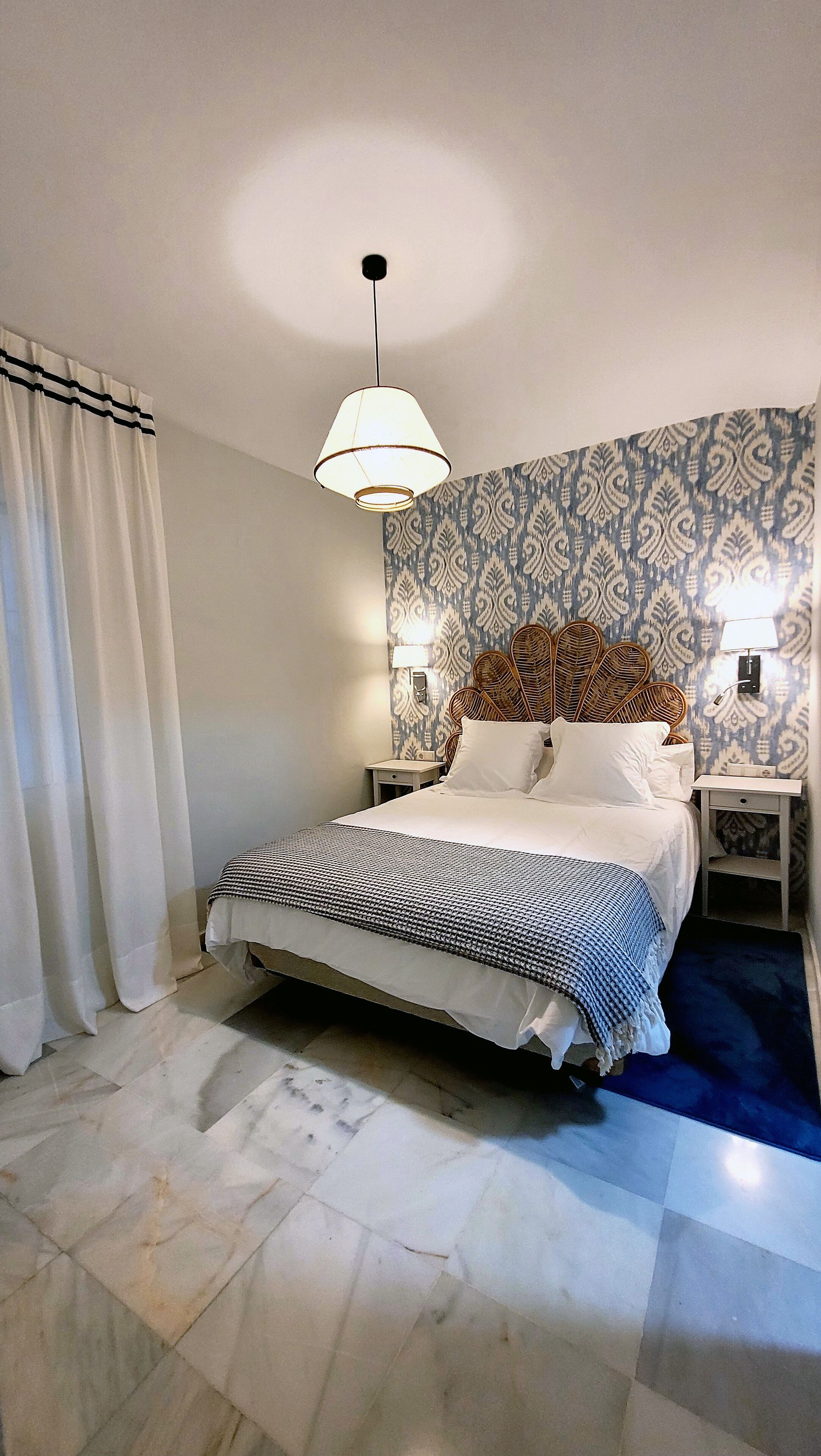 A bedroom with a bed, two bedside tables, a hanging light, patterned wallpaper, and a curtain.