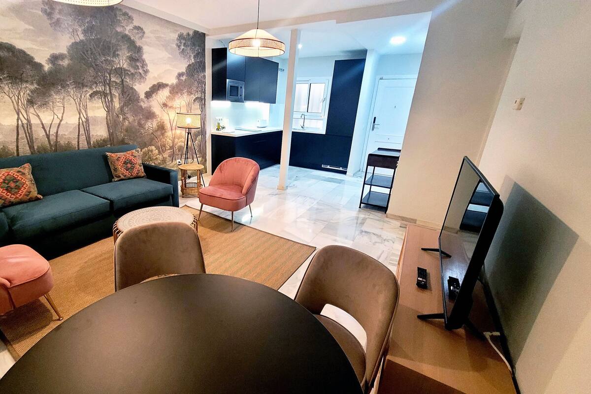 A living room with a sofa, chairs, a dining table, and a TV. There is a kitchen in the background.