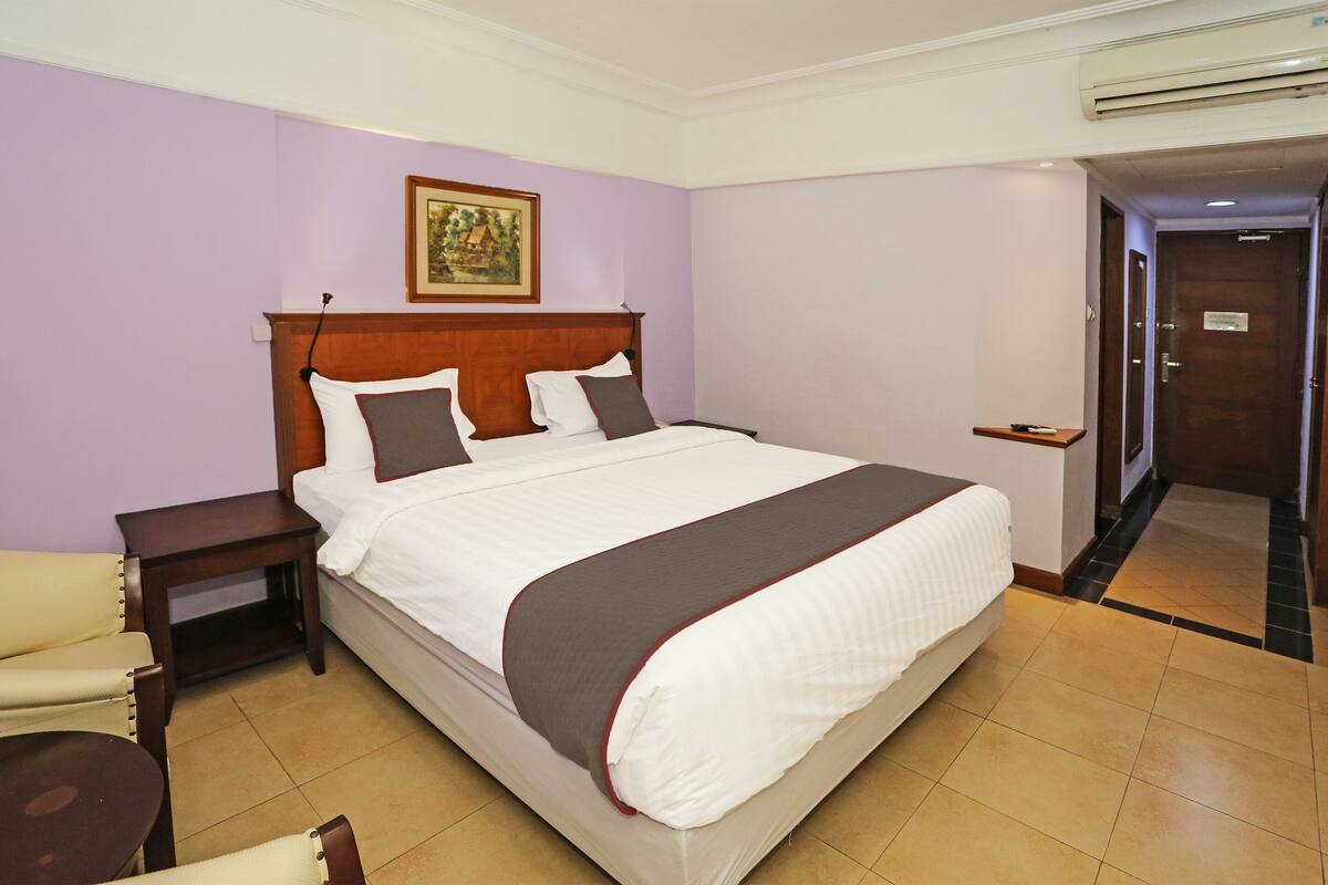 Ein Hotelzimmer mit einem großen Bett, einem Nachttisch, einem Sessel und einer Tür zu einem weiteren Raum.