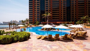 Zona de piscina en un resort con tumbonas y sombrillas, rodeada de palmeras y un edificio grande al fondo.