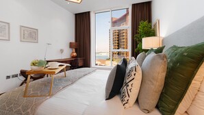 Una habitación de hotel moderna con una cama grande, una mesita de madera con un frutero, un escritorio con una lámpara y vistas a un edificio a través de un ventanal amplio.