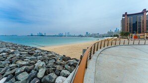 Un paisaje urbano costero con una playa arenosa, un rompeolas rocoso y un moderno edificio de gran altura al fondo.